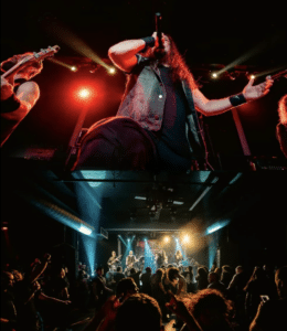 A rock band performs on stage under colorful lights, with a lead singer holding a microphone and a crowd watching in a dark concert venue.